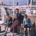 Colour photograph showing woman and two young boys sitting on a bench on the deck of a P&O ship, a lifeboat in view behind them. The woman wears a long tartan skirt, navy top and patterned scarf and looks directly into the camera. The two boys are dressed in shorts, blazers and long grey socks. The boy to her right is looking up from his book and the boy to her left is peeking up too, either shyly or squinting in the sunlight.