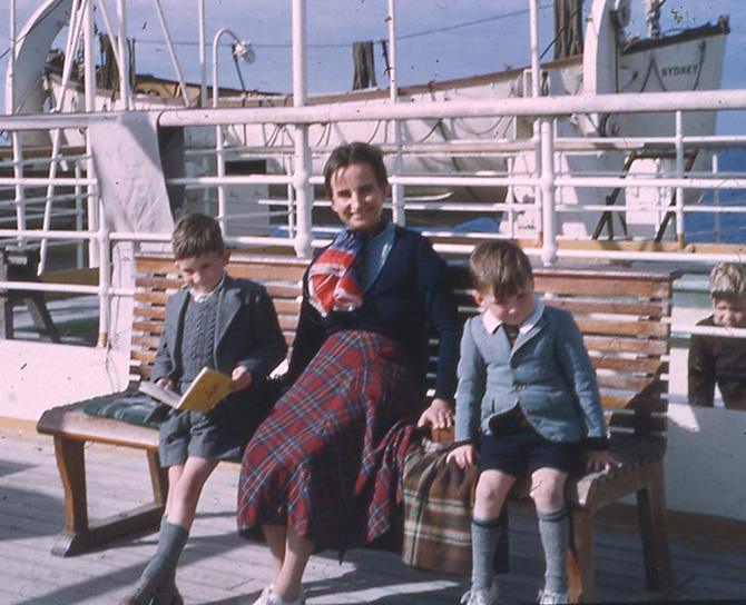 Colour photograph showing woman and two young boys sitting on a bench on the deck of a P&O ship, a lifeboat in view behind them. The woman wears a long tartan skirt, navy top and patterned scarf and looks directly into the camera. The two boys are dressed in shorts, blazers and long grey socks. The boy to her right is looking up from his book and the boy to her left is peeking up too, either shyly or squinting in the sunlight.