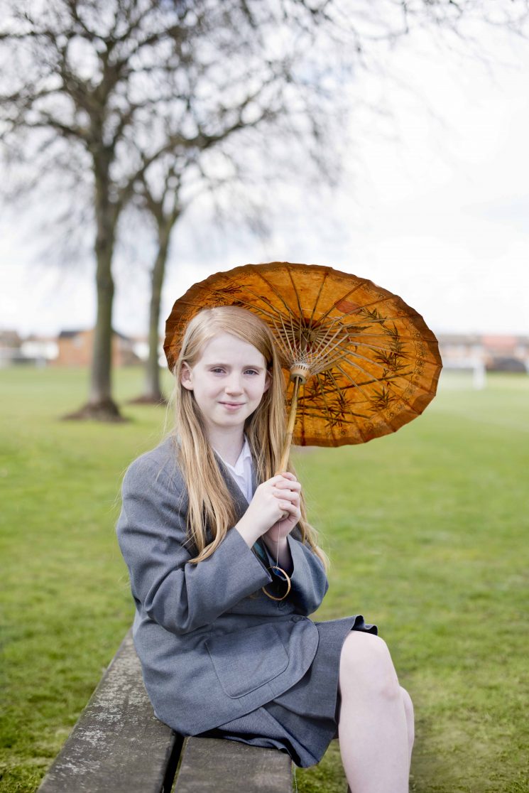 Pupil in uniform holds pretty autumnal-coloured parasol in park with trees behind.