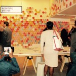 Visitors look at paper discs on wall, which visitors have written their migration/non-migration stories on before hanging up. A young boy in the foreground captures the scene on a tablet camera.