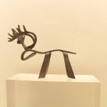 A small artistic elegant metal reindeer with a curved back and head, simple legs and intricate antlers on a small off-white podium with an off-white wall behind.