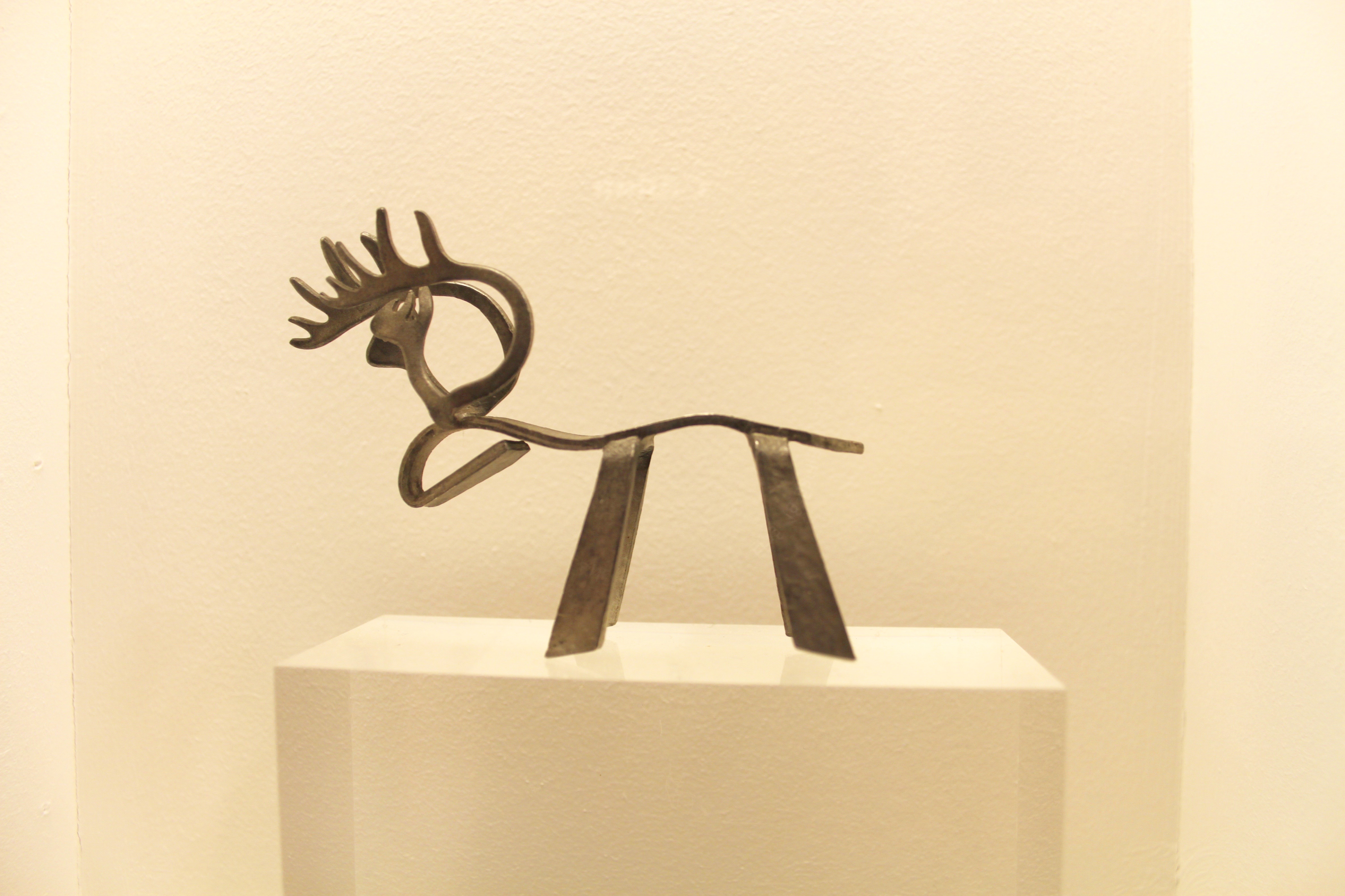 A small artistic elegant metal reindeer with a curved back and head, simple legs and intricate antlers on a small off-white podium with an off-white wall behind.